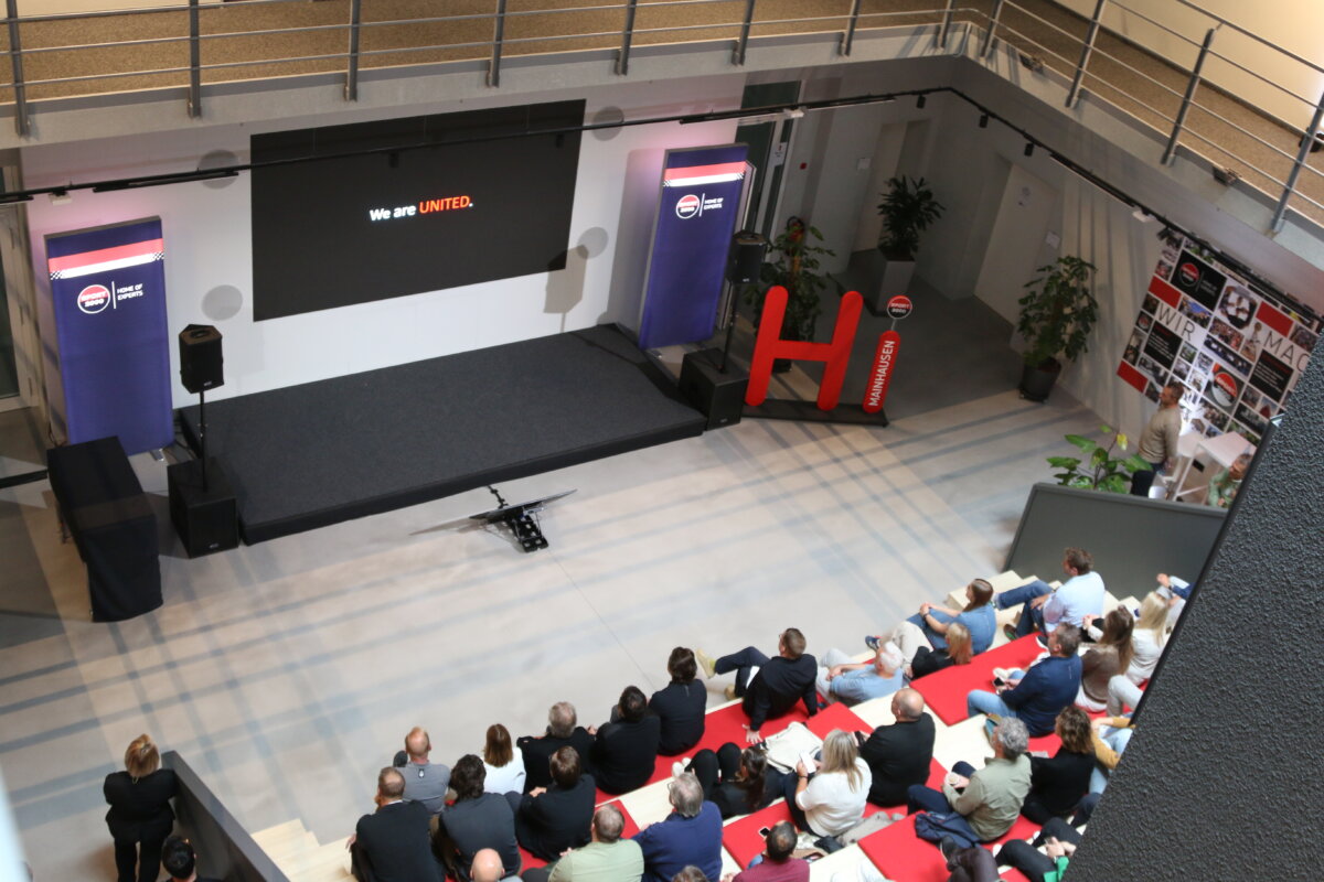 In front of a modern presentation area, there is a tiered grandstand filled with numerous people. The attendees are watching the presentation attentively and directing their attention toward the stage.