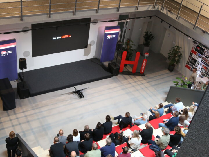 In front of a modern presentation area, there is a tiered grandstand filled with numerous people. The attendees are watching the presentation attentively and directing their attention toward the stage.